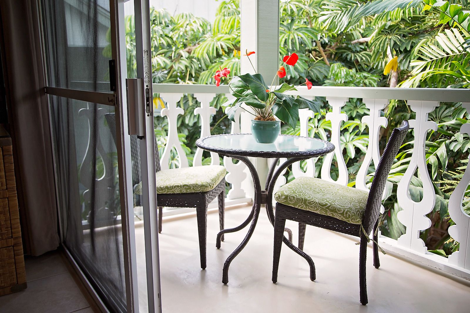 A balcony with a glass door, two wicker chairs with patterned cushions, a small round table, and a potted plant with red flowers.