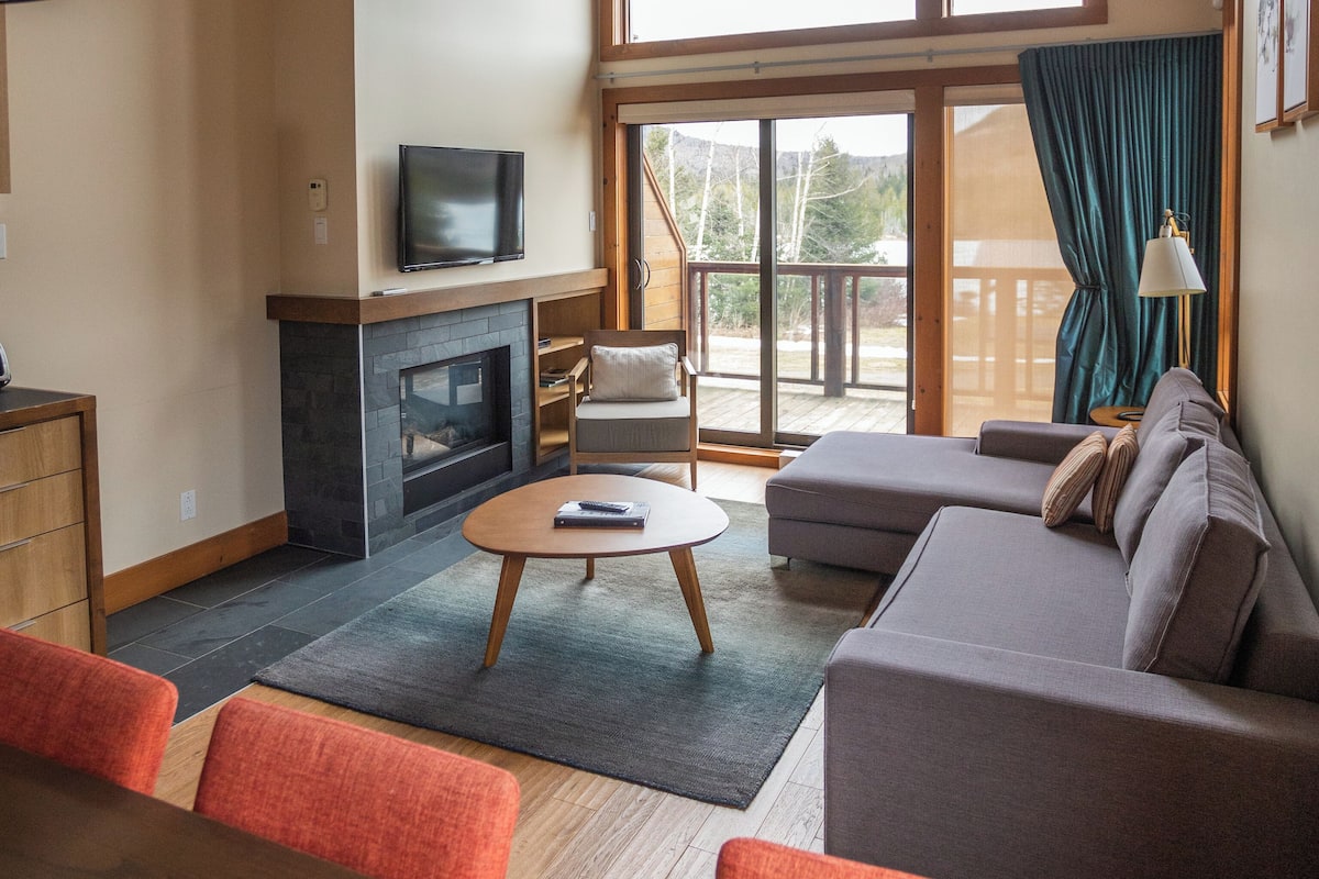 A spacious living room with a fireplace, a large window, and a balcony.
