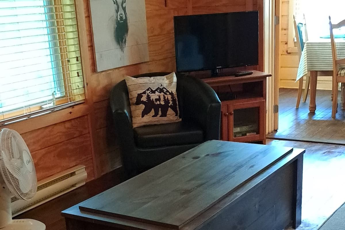 A living room with a wooden floor, a dark wooden coffee table, a black leather sofa with a patterned pillow, a television on a wooden stand, and a fan.