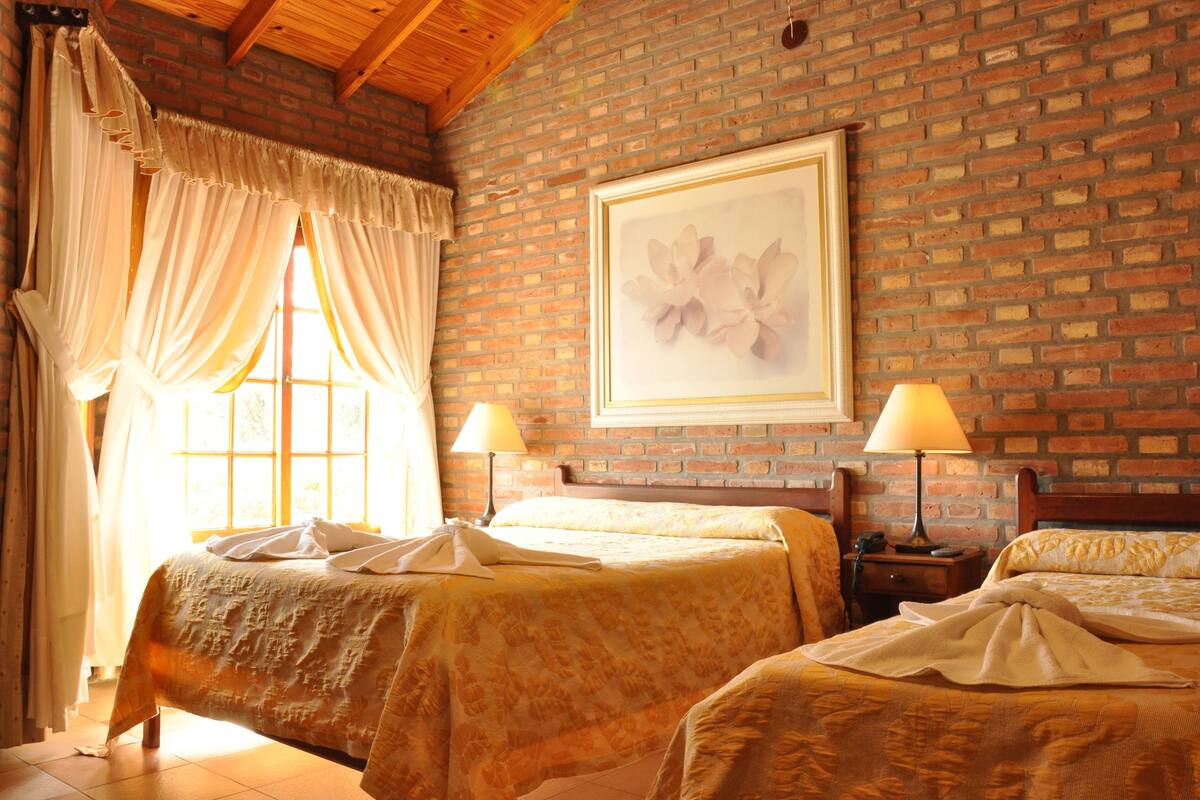 Ein Hotelzimmer mit zwei Betten, einem großen Fenster mit Vorhängen, einem gerahmten Bild an der Wand und einer Ziegelwand.