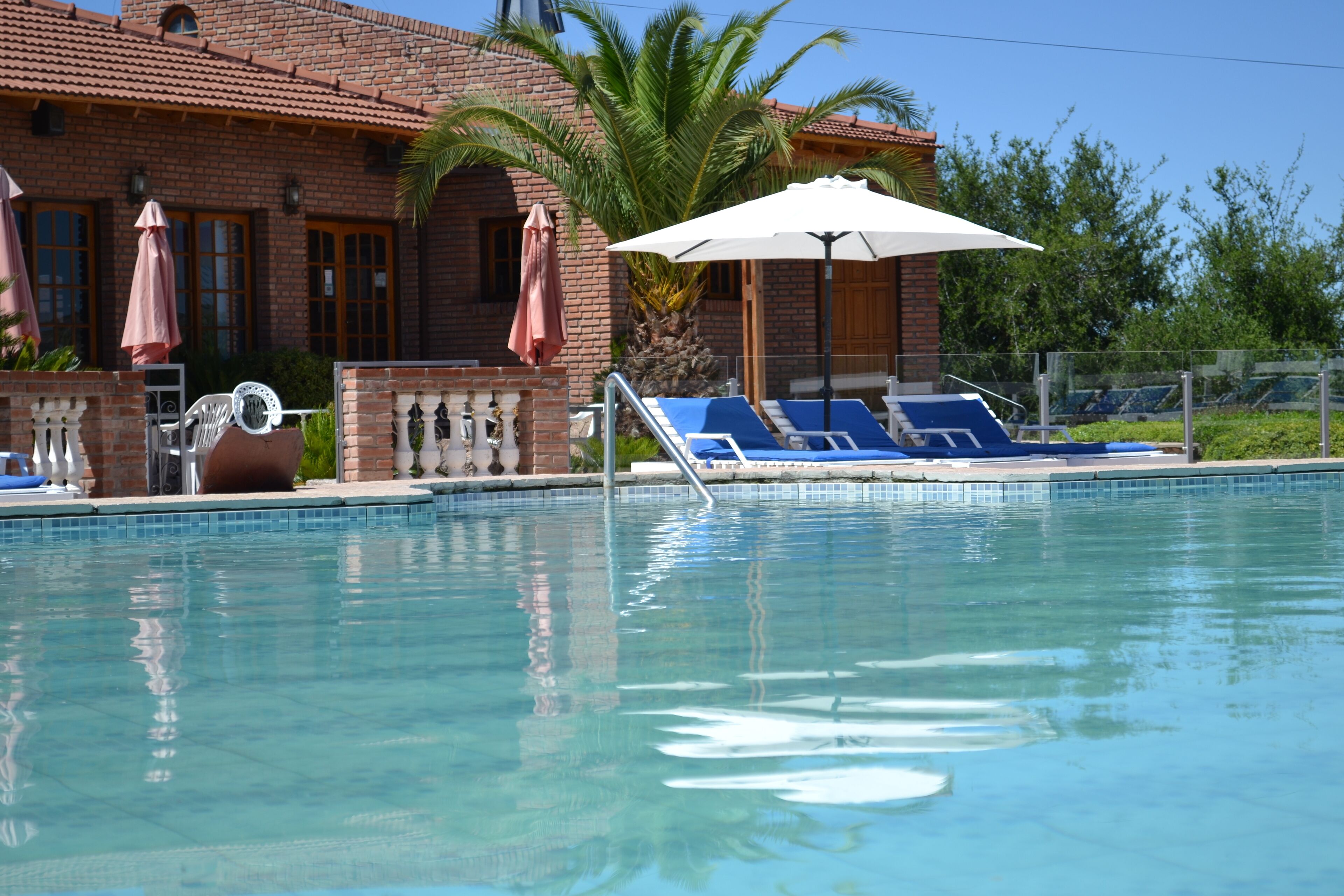 A swimming pool with lounge chairs and umbrellas, a brick building with a tiled roof, and palm trees in the background.