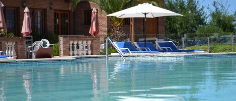 Una piscina con sillones de playa y sombrillas, un edificio de ladrillo con tejado de tejas, y palmeras al fondo.