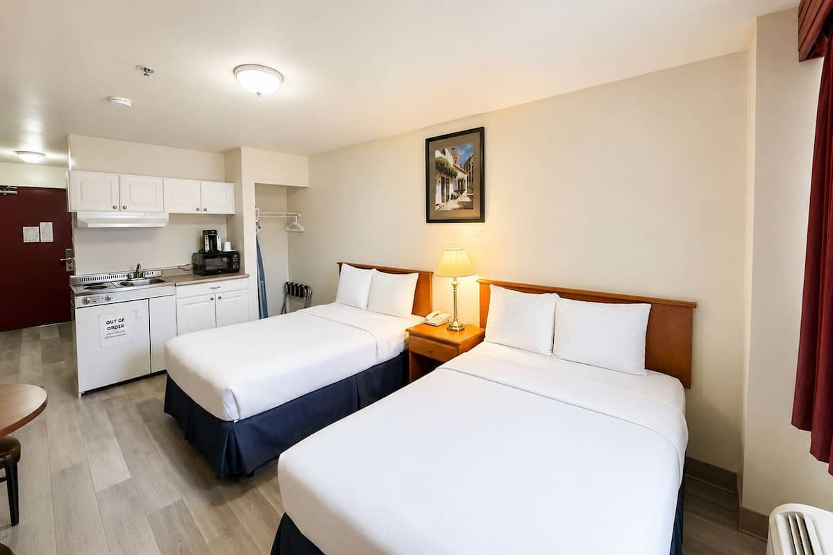A hotel room with two beds, a kitchenette, a painting on the wall, and a window with curtains.