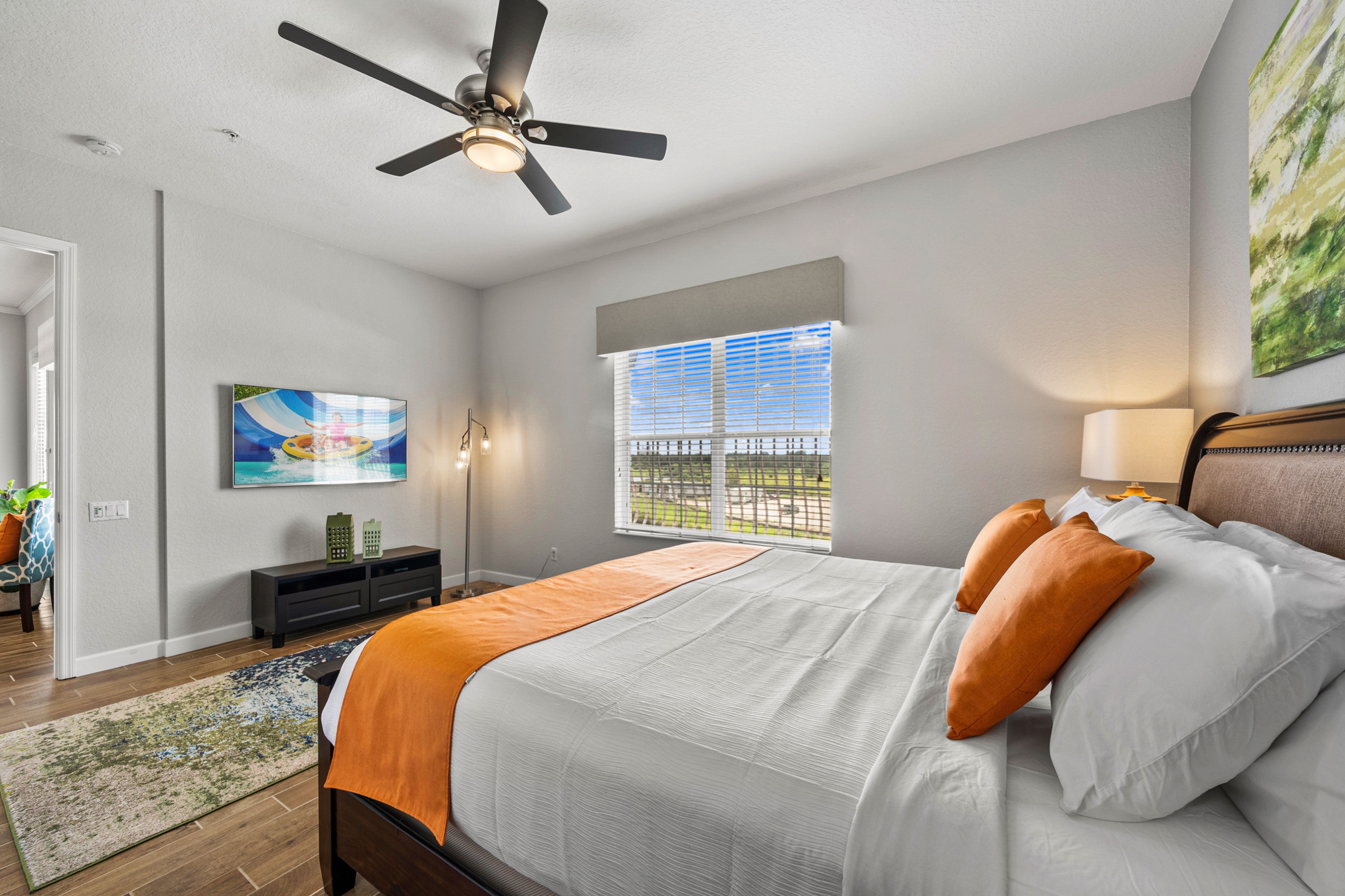 A bedroom with a bed, a ceiling fan, a window with blinds, a painting on the wall, and a nightstand with a lamp.