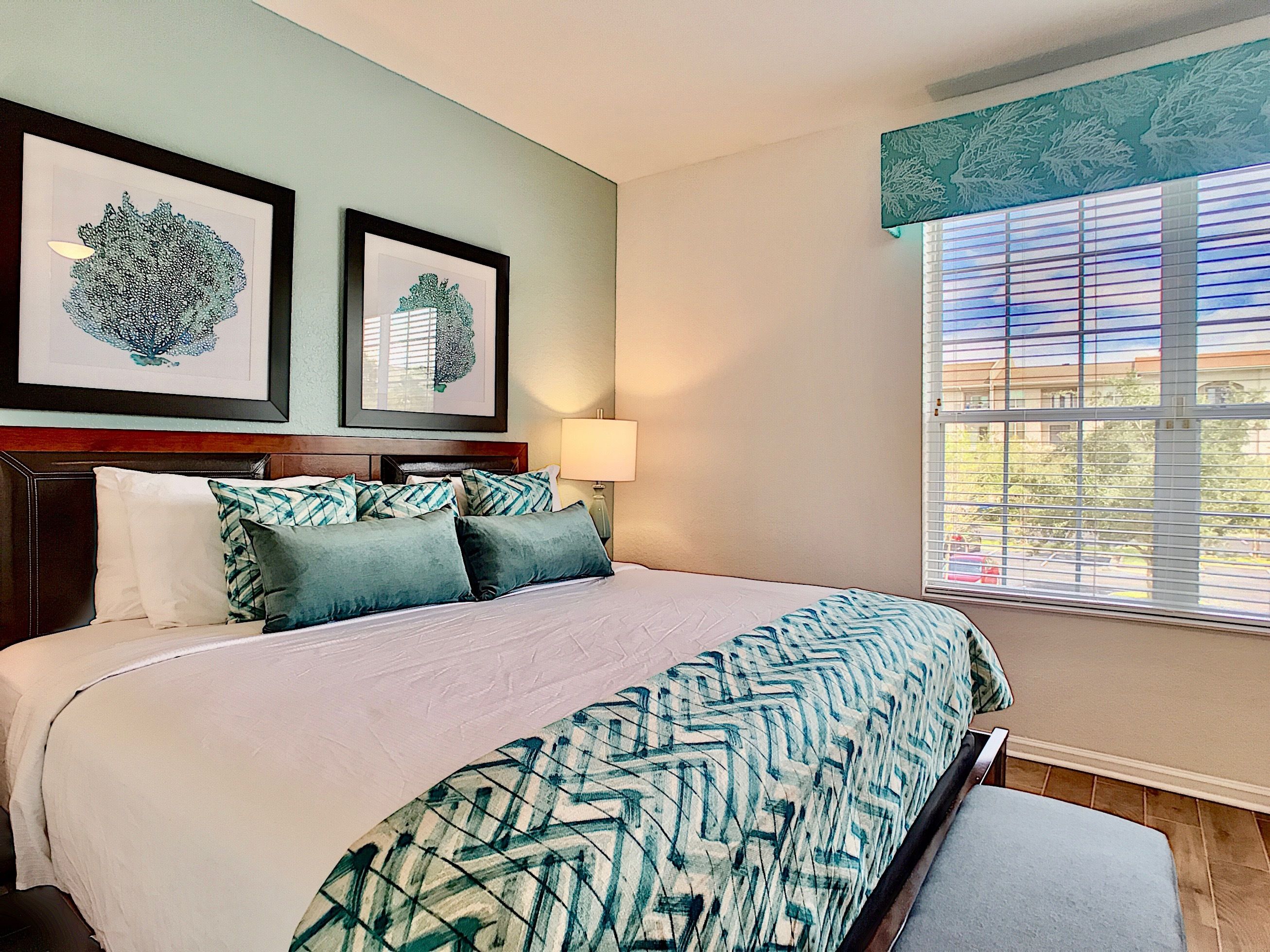 A hotel room with a large bed, two framed artworks on the wall, a window with blinds, and a wooden headboard.