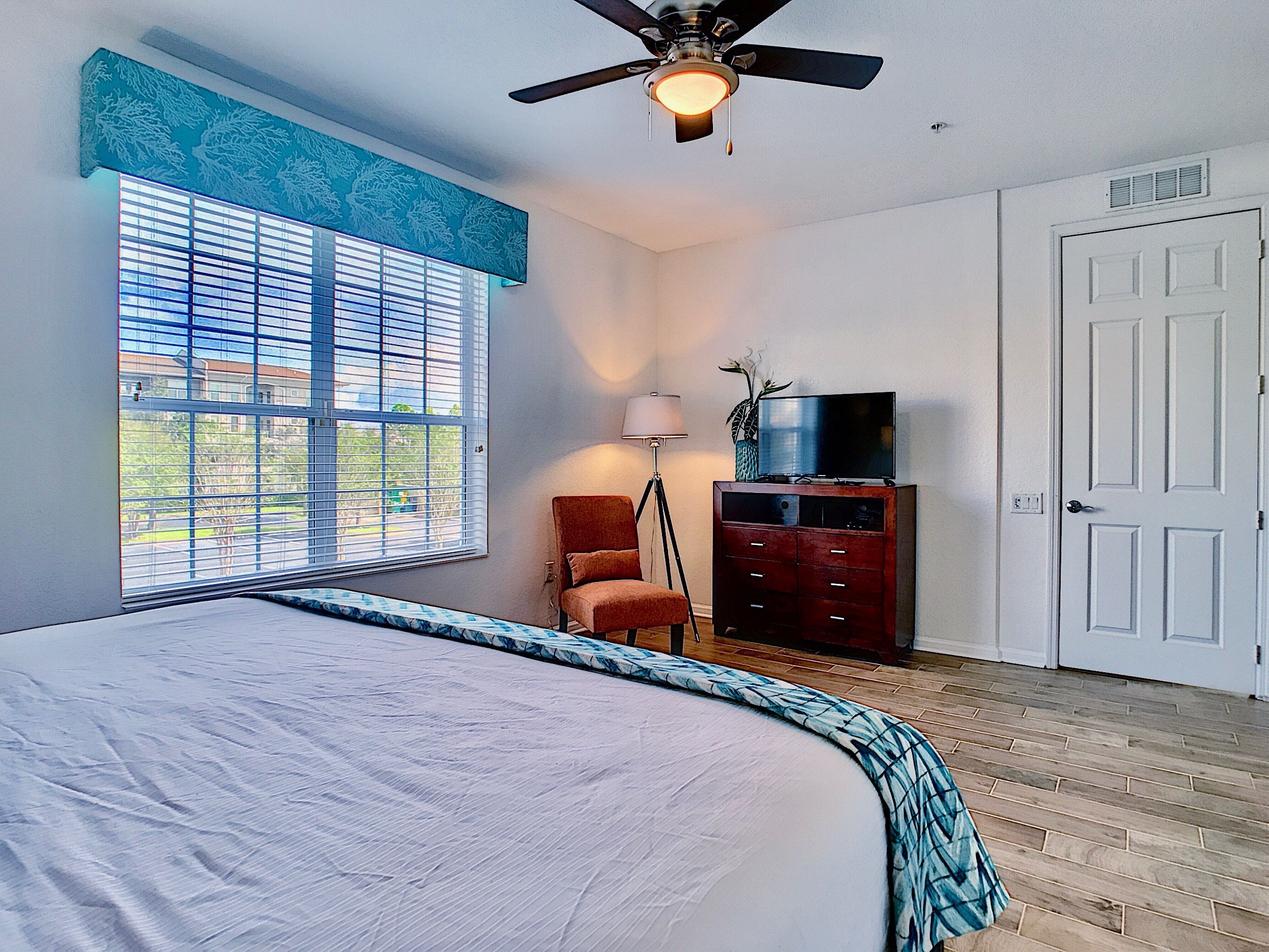 Une chambre à coucher avec un grand lit, un ventilateur de plafond, une commode, un téléviseur et une fenêtre avec des stores.