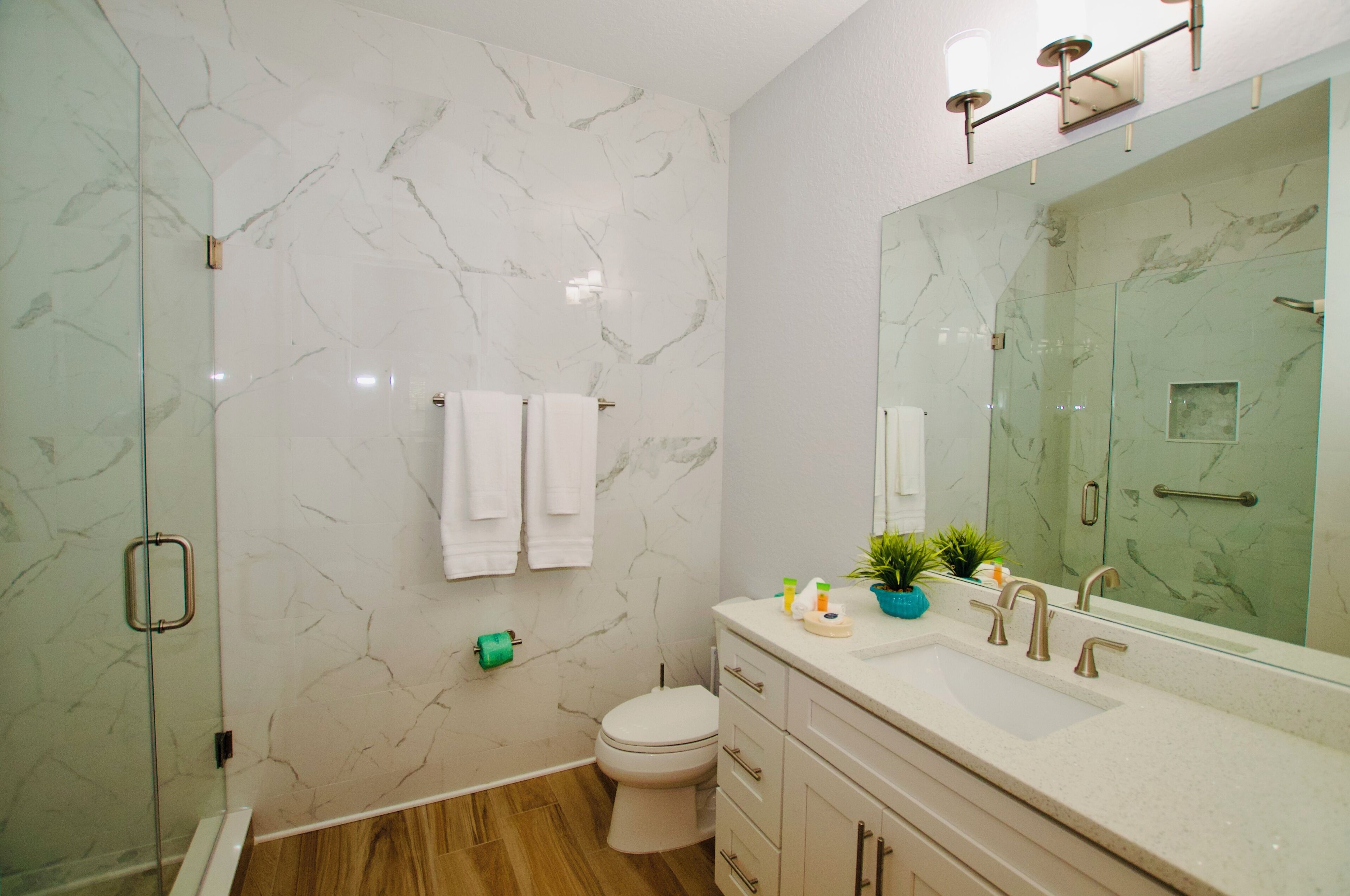 A modern bathroom with a marble wall, a glass shower enclosure, a white toilet, a vanity with a sink, and a large mirror.
