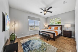A bedroom with a bed, a ceiling fan, a window with blinds, a nightstand, a lamp, a bench, and a rug.