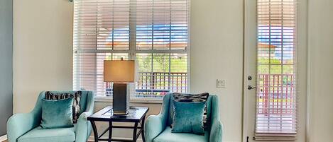 A living room with two light blue armchairs, a small black coffee table, and a floor lamp. There is a large window with blinds, a door with a red doormat, and a colorful abstract painting above the window.