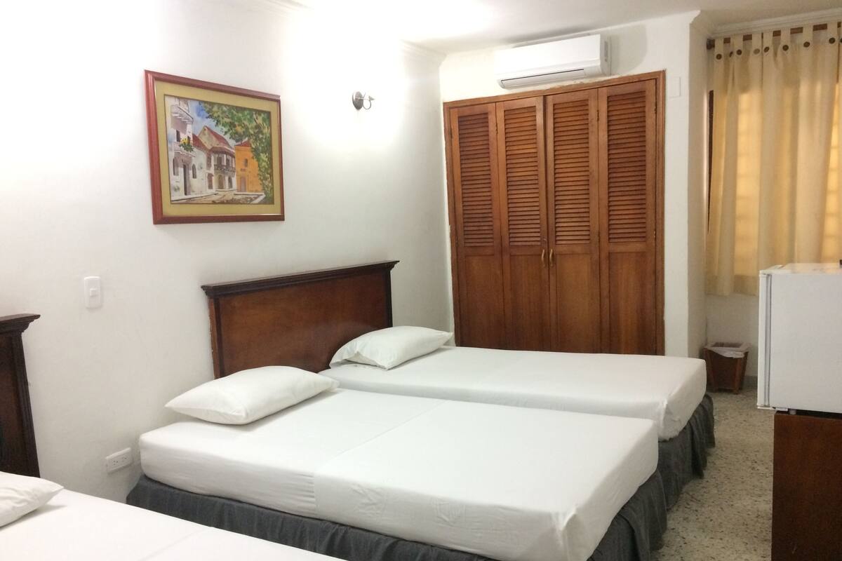 A hotel room with two single beds, a wooden wardrobe, a small refrigerator, and a painting on the wall.