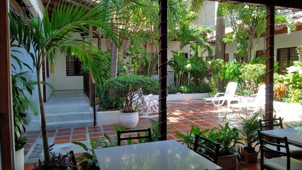 Uma área de estar externa com piso de azulejos, cadeiras de lounge brancas e plantas em vasos.
