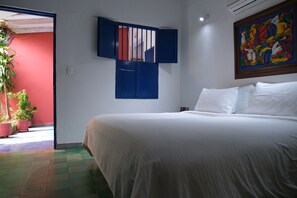 Une chambre avec un lit, une fenêtre avec des volets bleus, un tableau au mur et des plantes en pot.