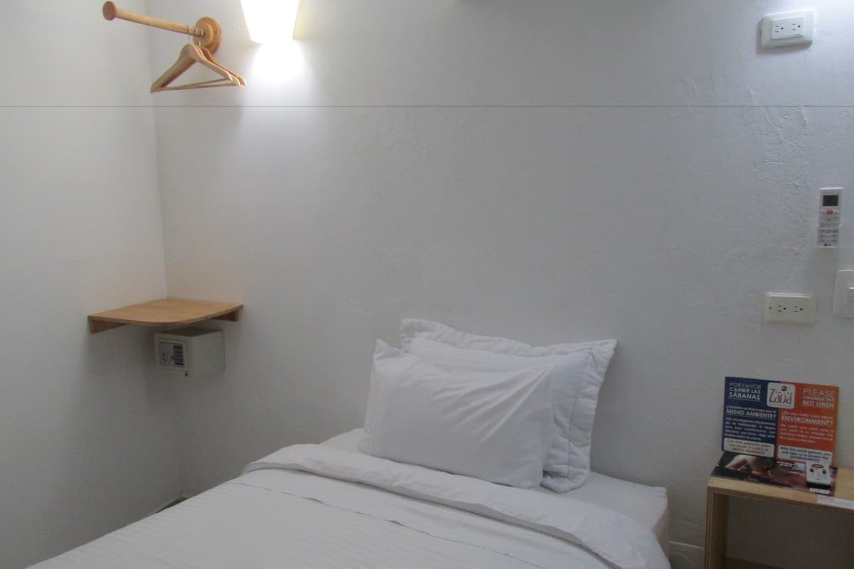 Ein kleines, weißes Zimmer mit einem Einzelbett, einem Holzregal, einer Steckdose und einem Hinweis zum Wechseln der Bettwäsche.