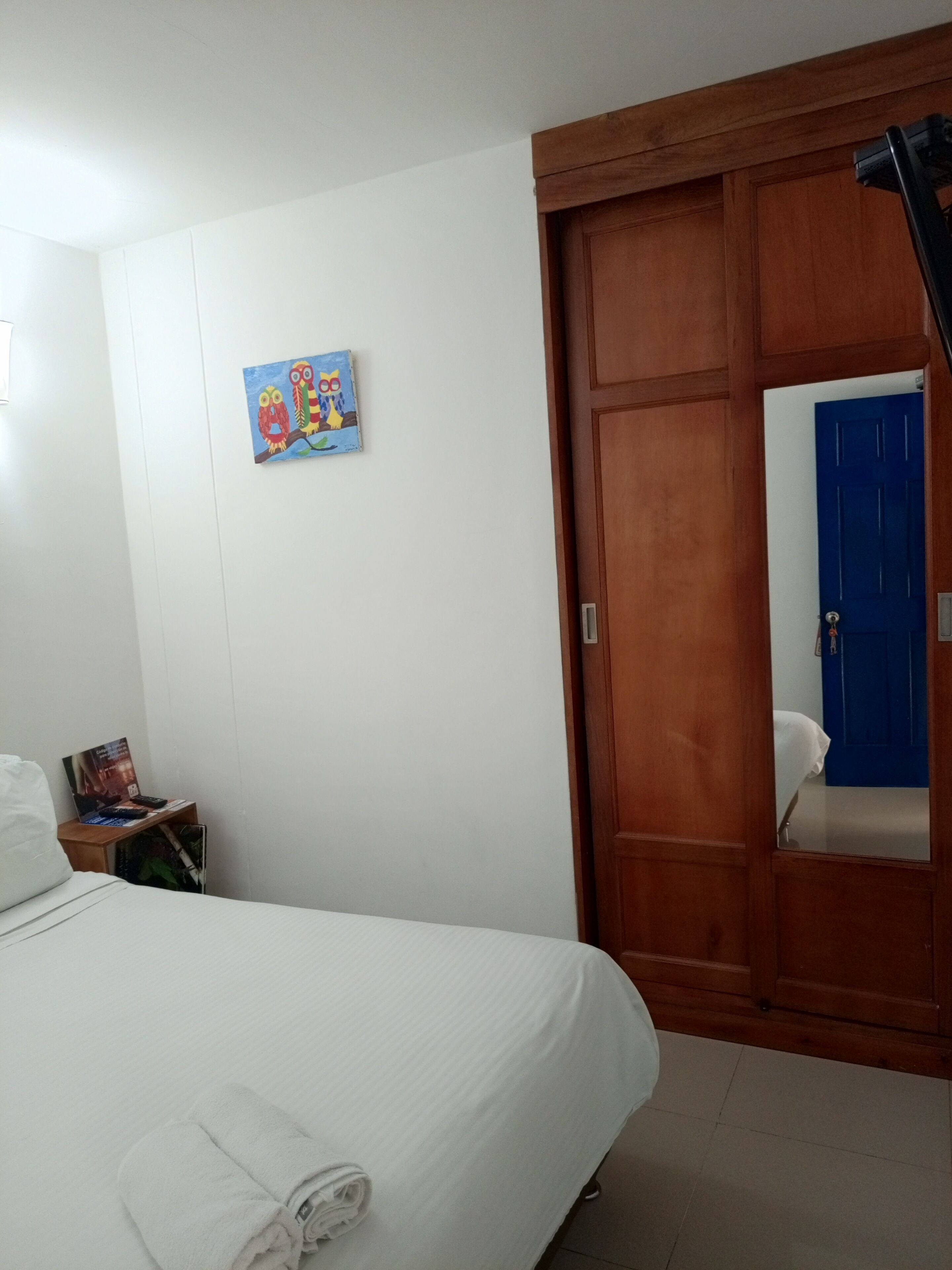 Une chambre d’hôtel avec un lit, une armoire en bois, une porte bleue et une petite table sur laquelle est posée une affiche.