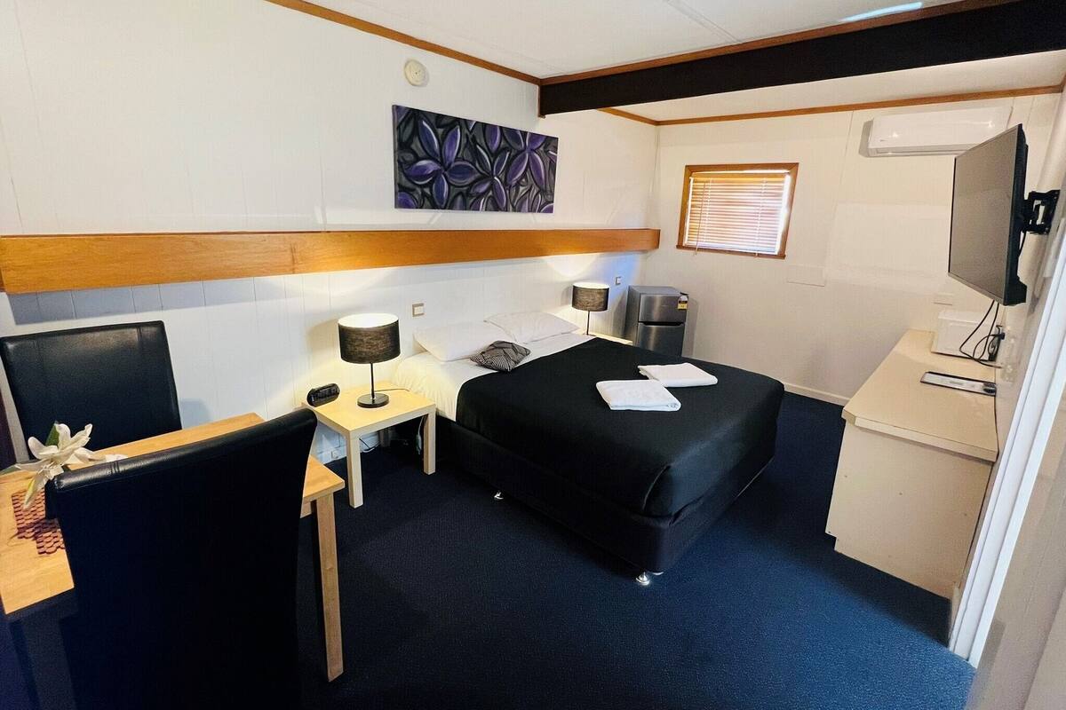 A hotel room with a bed, a desk, a chair, a small table, a lamp, a refrigerator, a TV, and a painting on the wall.