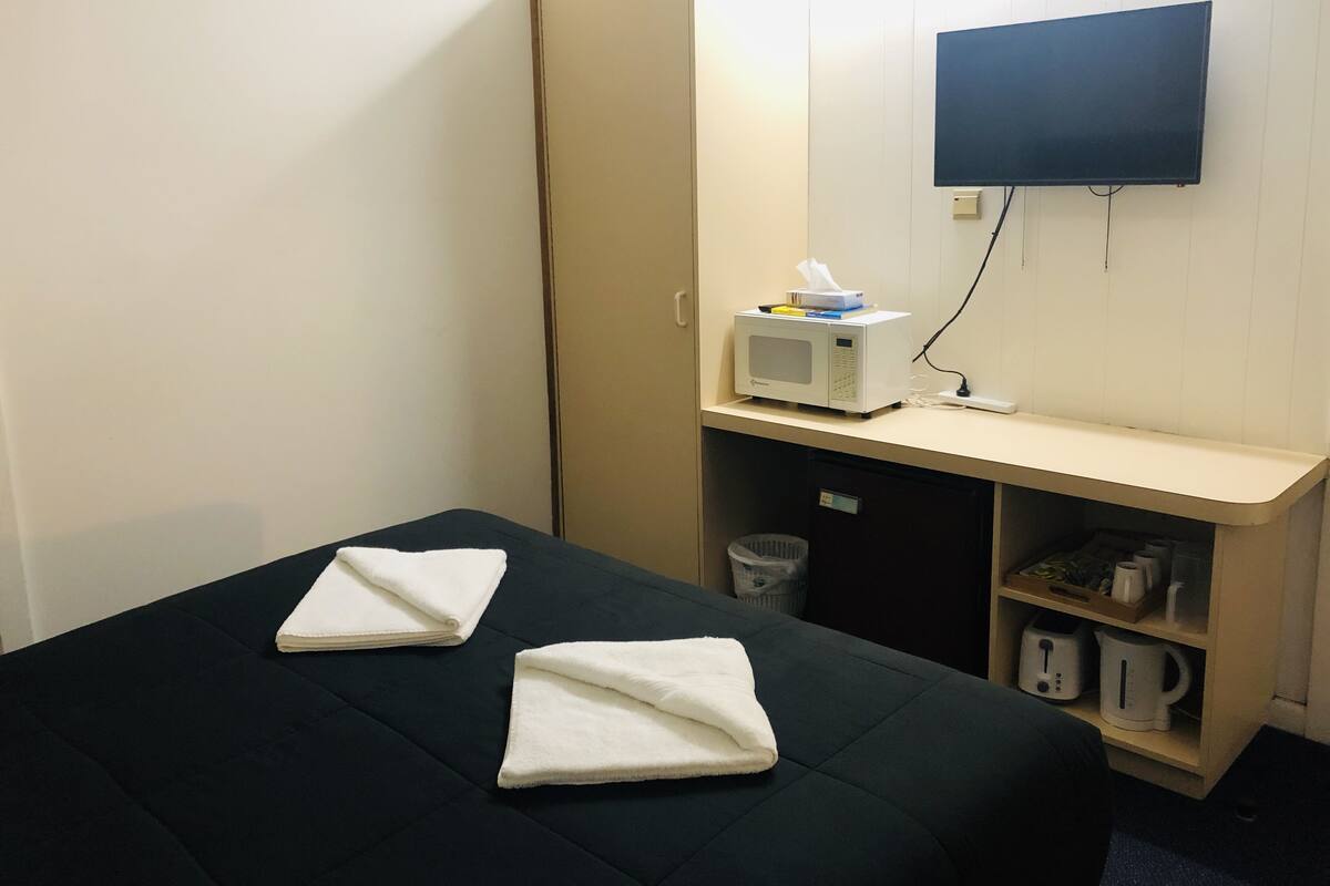 A hotel room with a bed, a TV mounted on the wall, a microwave on a small table, and a shelf with kitchen items.