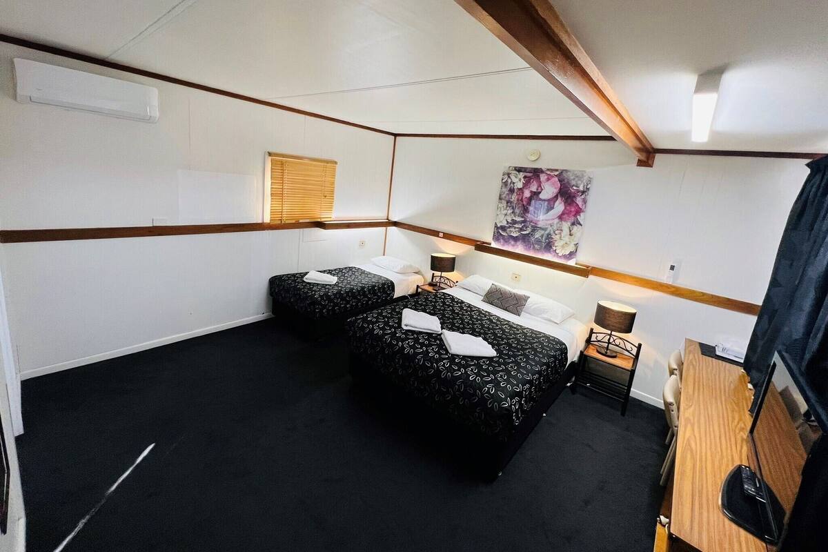 A hotel room with two beds, a wooden ceiling, a window with blinds, a painting on the wall, and a desk with a chair.