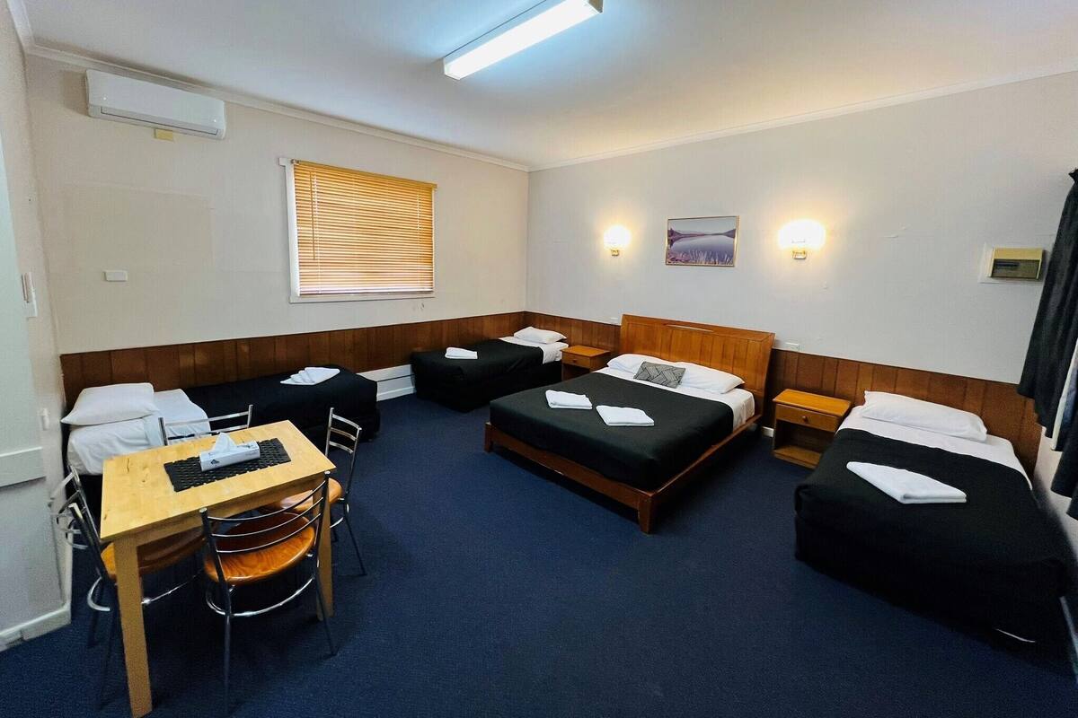 A hotel room with a single bed, two single beds, a desk with a chair, and a small table.
