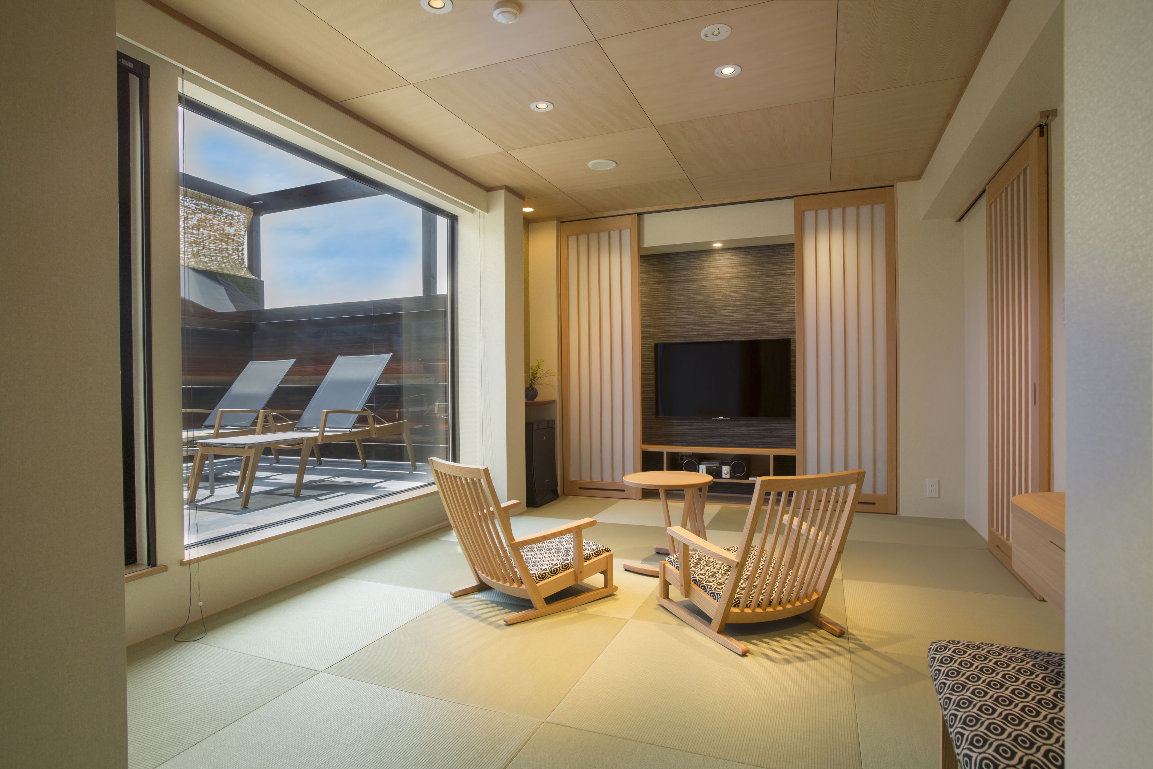 A modern living room with tatami flooring, a TV, and a seating area with two wooden chairs.