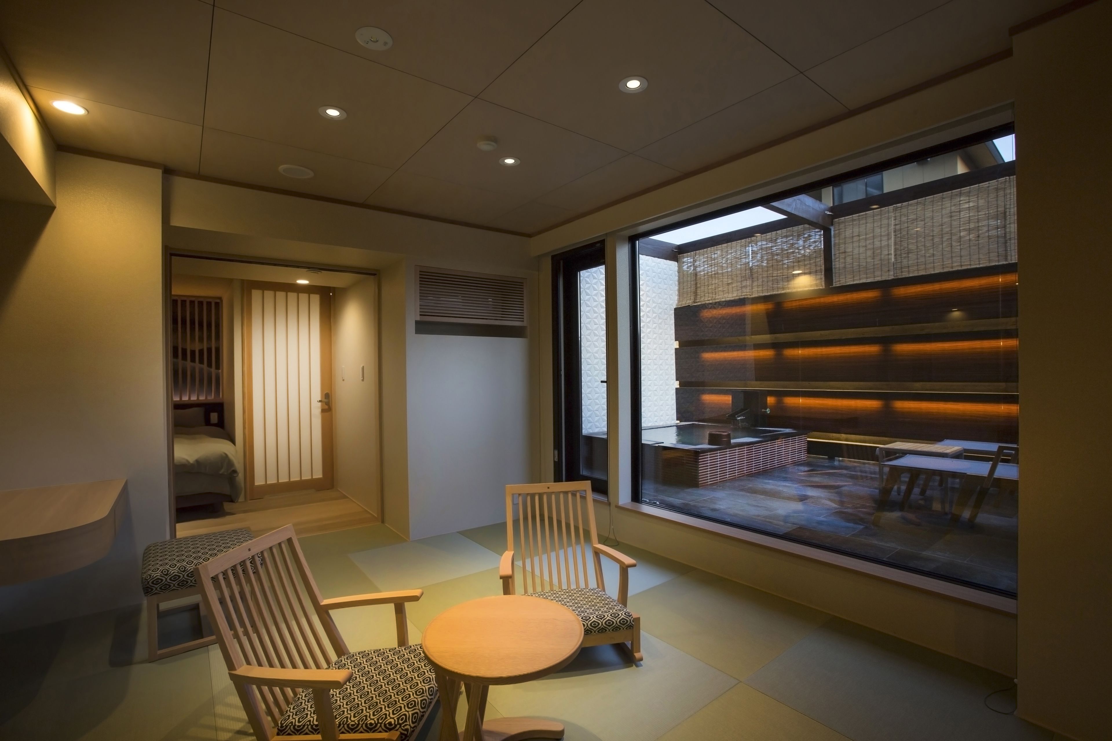 A modern interior with a seating area featuring two wooden chairs and a small round table, a large window with a view of an outdoor seating area, and a door leading to a room with a bed.