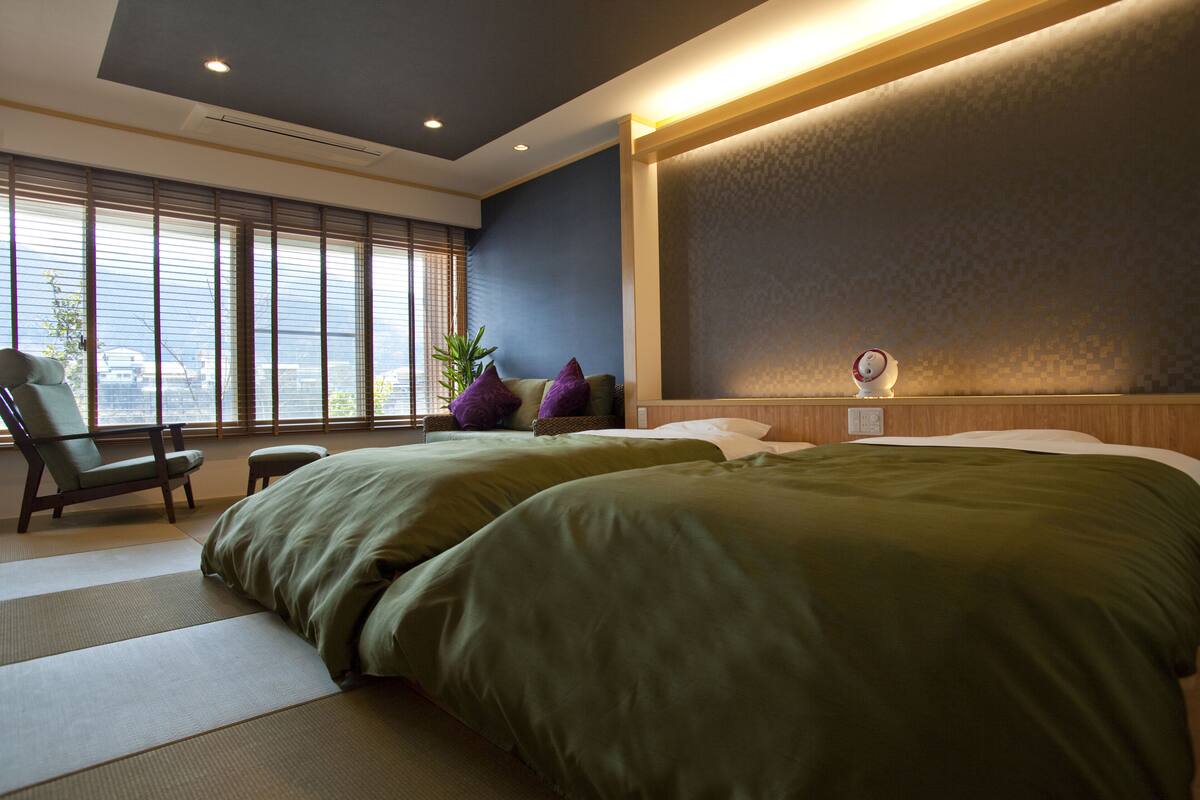 Een hotelkamer met twee bedden, een stoel, een klein tafeltje en een groot raam met zonwering.
