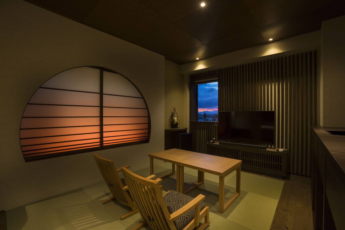 Een moderne woonkamer met een televisie, een eettafel en een groot raam met uitzicht op een zonsondergang.