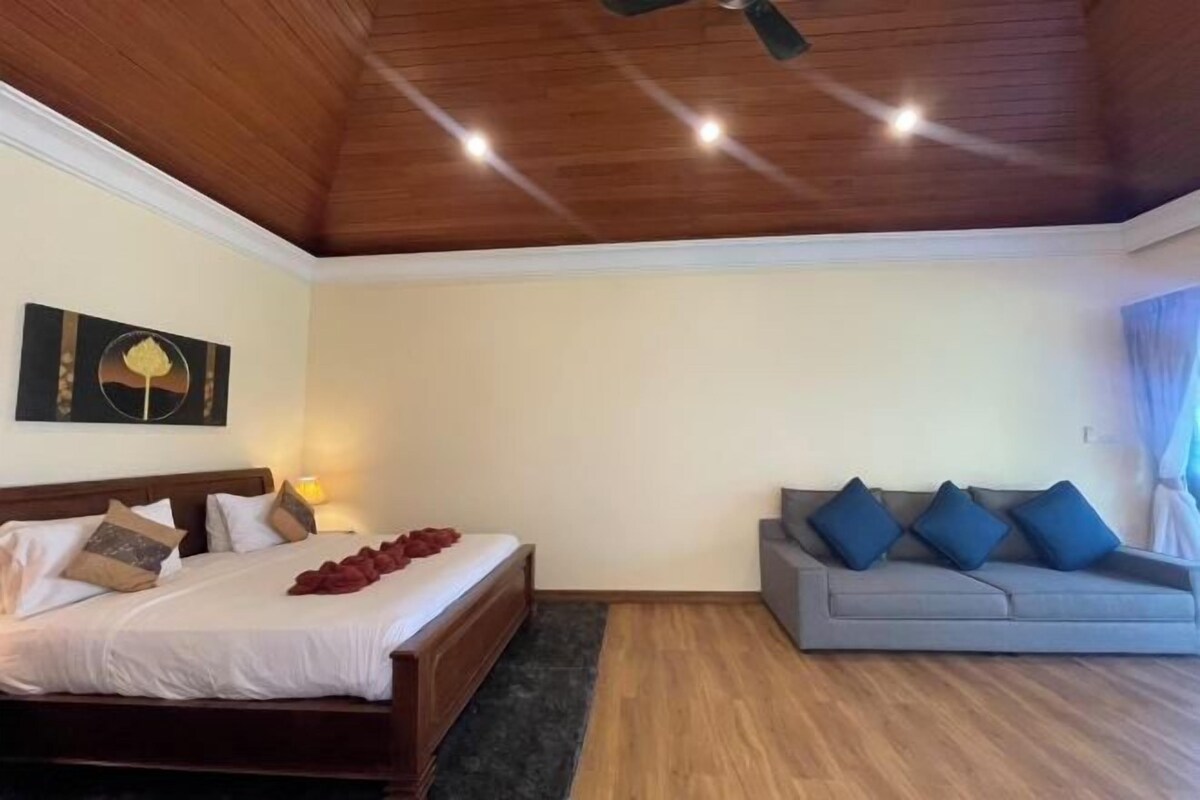 Een slaapkamer met een houten plafond, een bed met kussens, een grijze bank en een wandlamp.