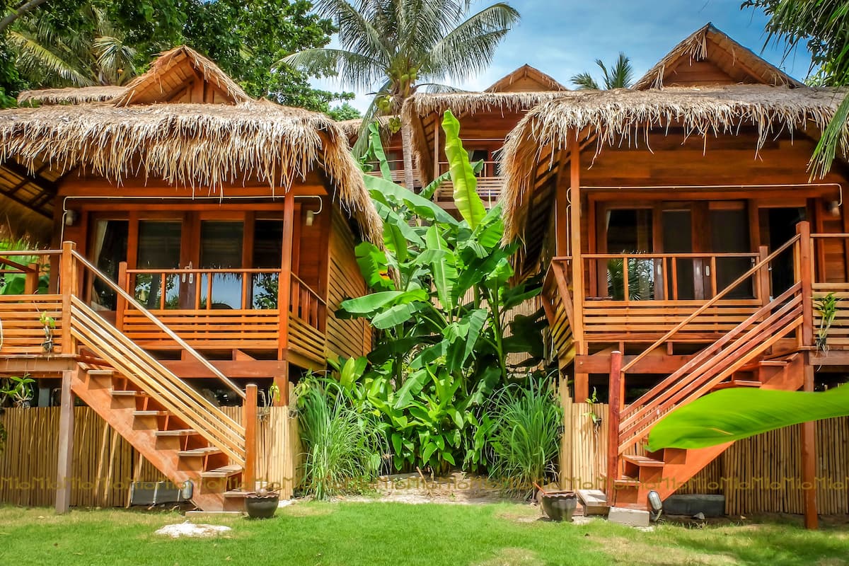 Two wooden bungalows with thatched roofs, surrounded by lush greenery.