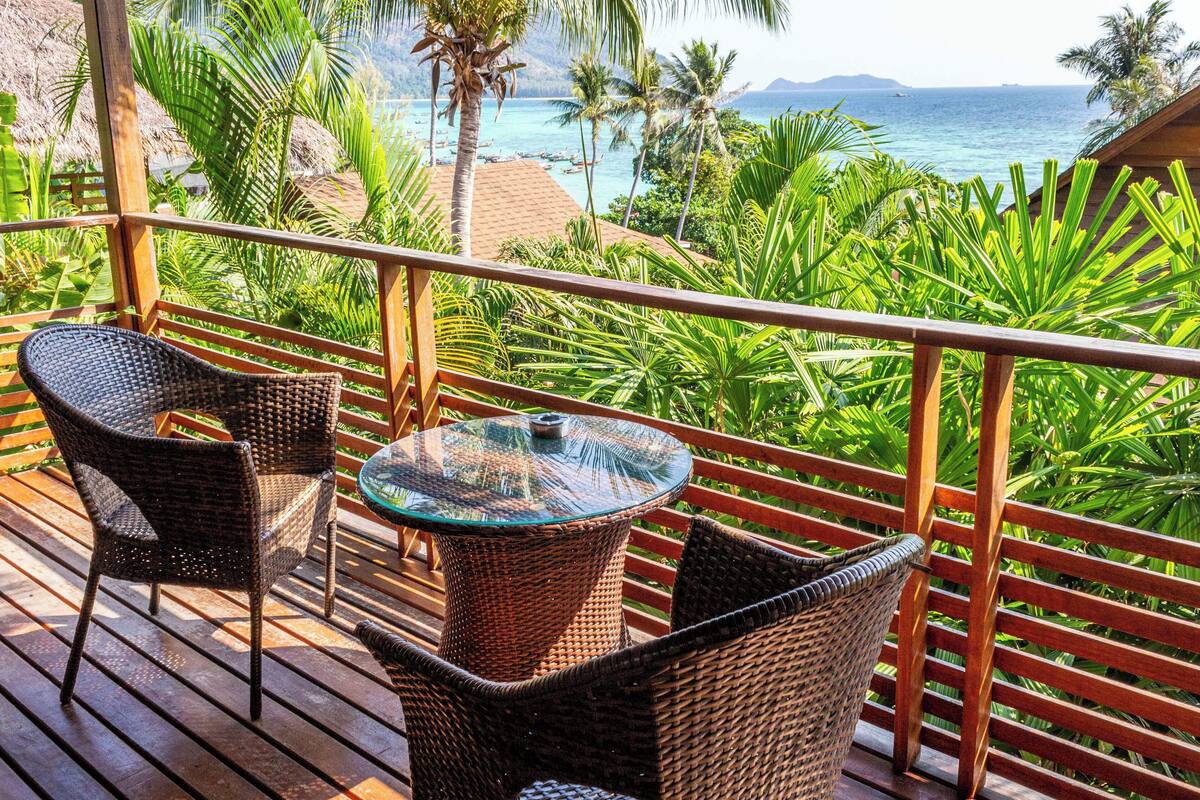 A wooden deck with wicker chairs and a glass table overlooking a beach with palm trees and the ocean.
