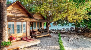 A wooden cabin with a balcony overlooking a beach, furnished with seating and a table.