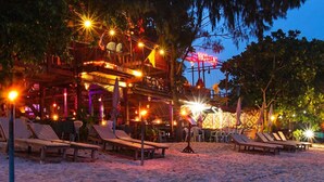 Un bar en la playa con sillones y sombrillas, rodeado de árboles e iluminado con luz cálida.