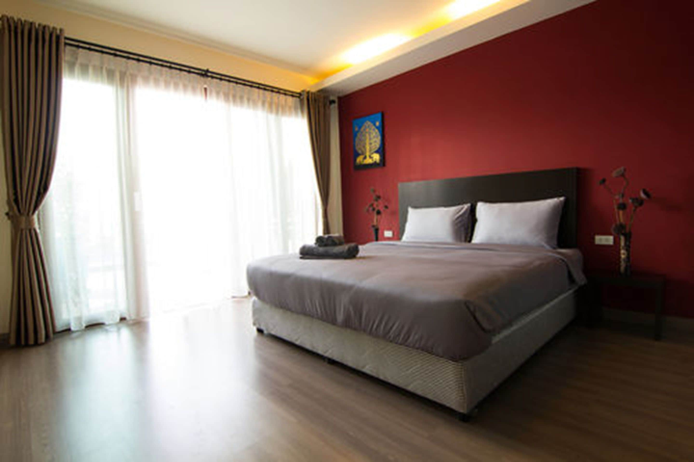 A modern hotel room with a large bed, a red accent wall, and a large window with curtains.