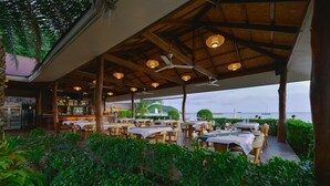 An outdoor dining area with tables and chairs, a covered roof, and a view of the sea.