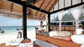 A beachside room with a bed, a window overlooking the sea, and a thatched roof.