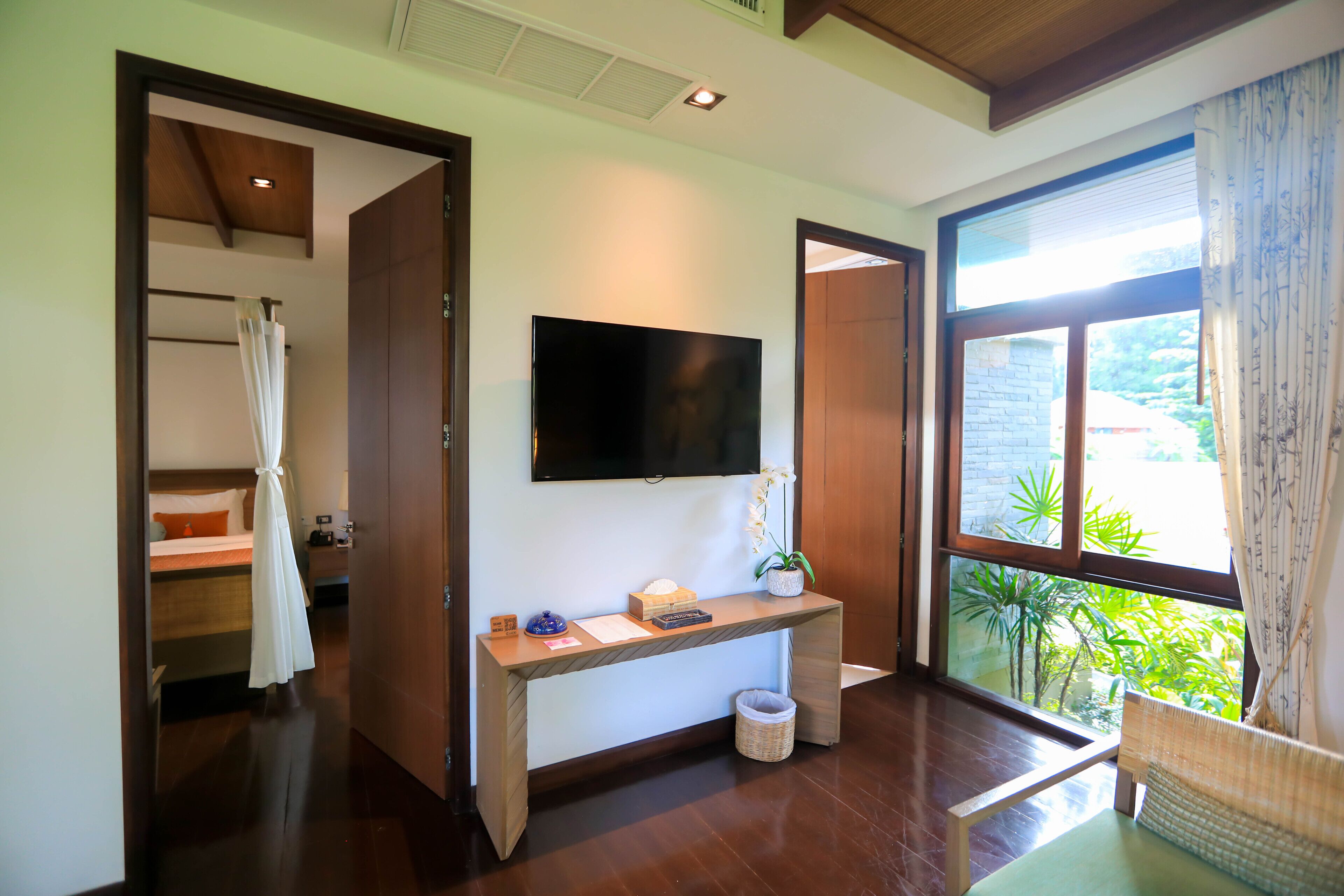 Une chambre d’hôtel avec un plancher en bois, un téléviseur fixé au mur, un bureau avec un panier, et une fenêtre donnant sur un paysage verdoyant.