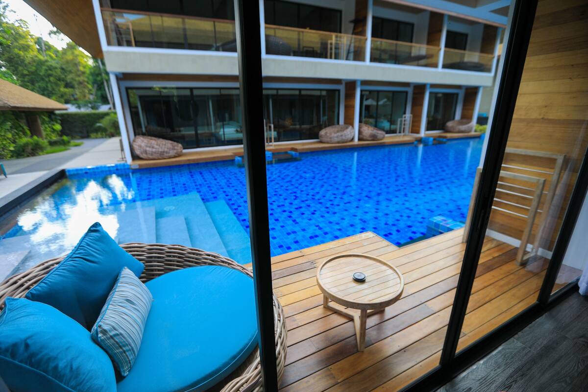 Área da piscina com uma espreguiçadeira de vime, uma pequena mesa de madeira e vista para um edifício alto com varandas.