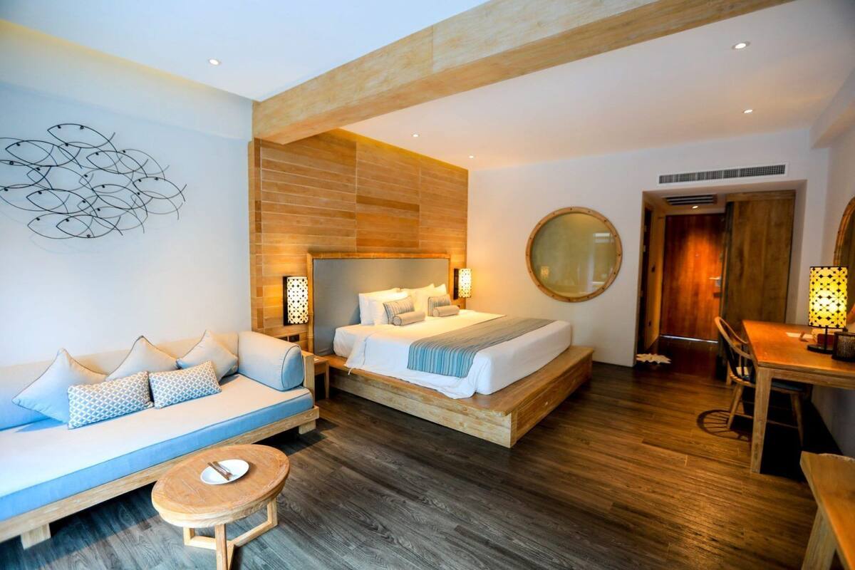 Quarto de hotel com cama, sofá, espelho redondo, mesa de jantar e uma pequena mesa redonda com uma xícara.
