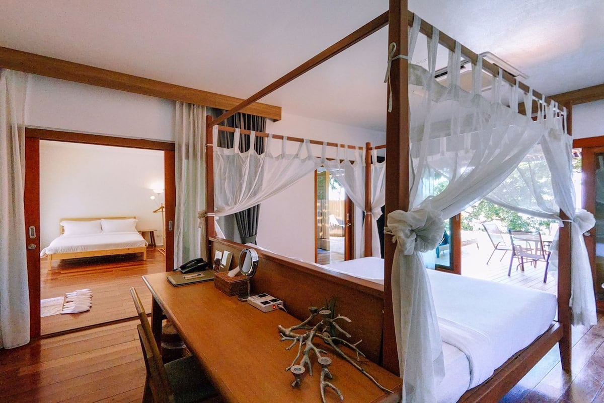 Una cama con dosel y cuatro columnas, un escritorio de madera con espejo, y vistas a un balcón con mobiliario exterior.