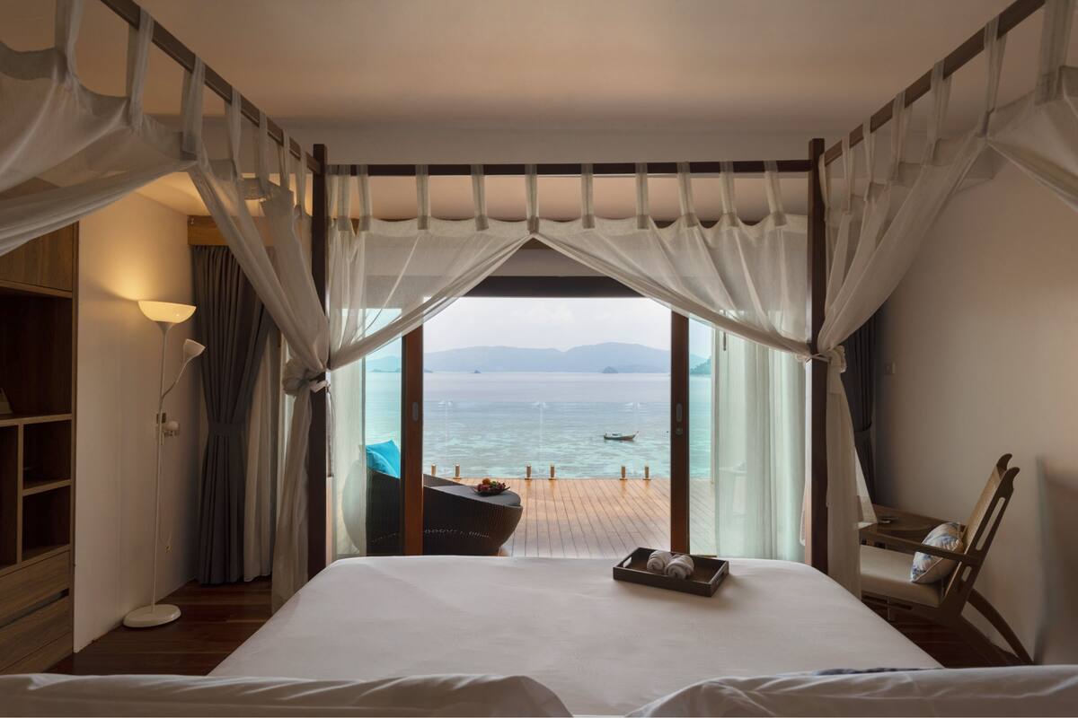 Un dormitorio con una cama grande, piso de madera, mesita de noche, una silla y vistas al mar a través de cortinas traslúcidas.