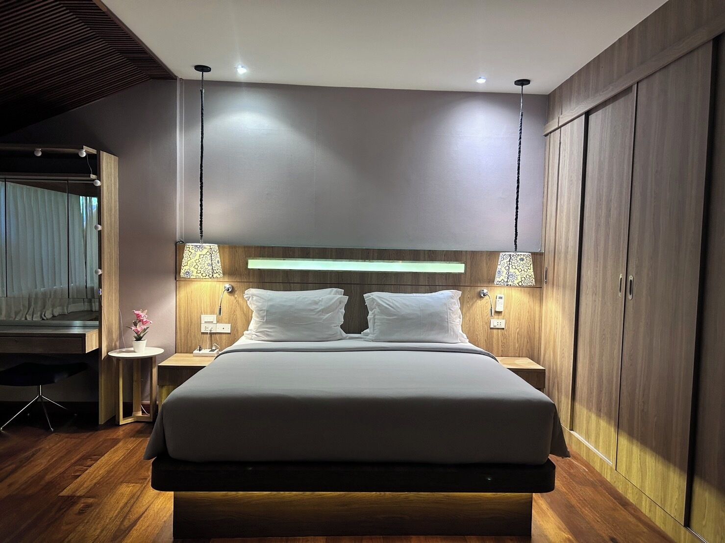 A modern hotel room with a large bed, wooden headboard, bedside lamps, and a wardrobe.