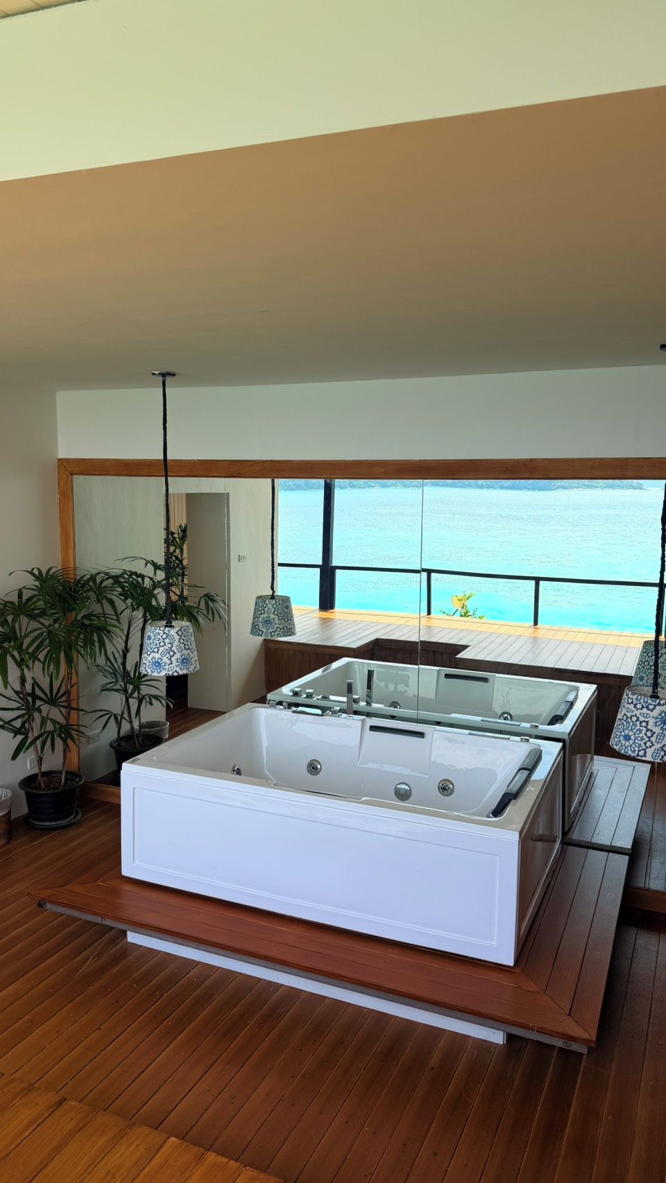 A modern bathroom with a large bathtub, wooden flooring, and a view of the sea.