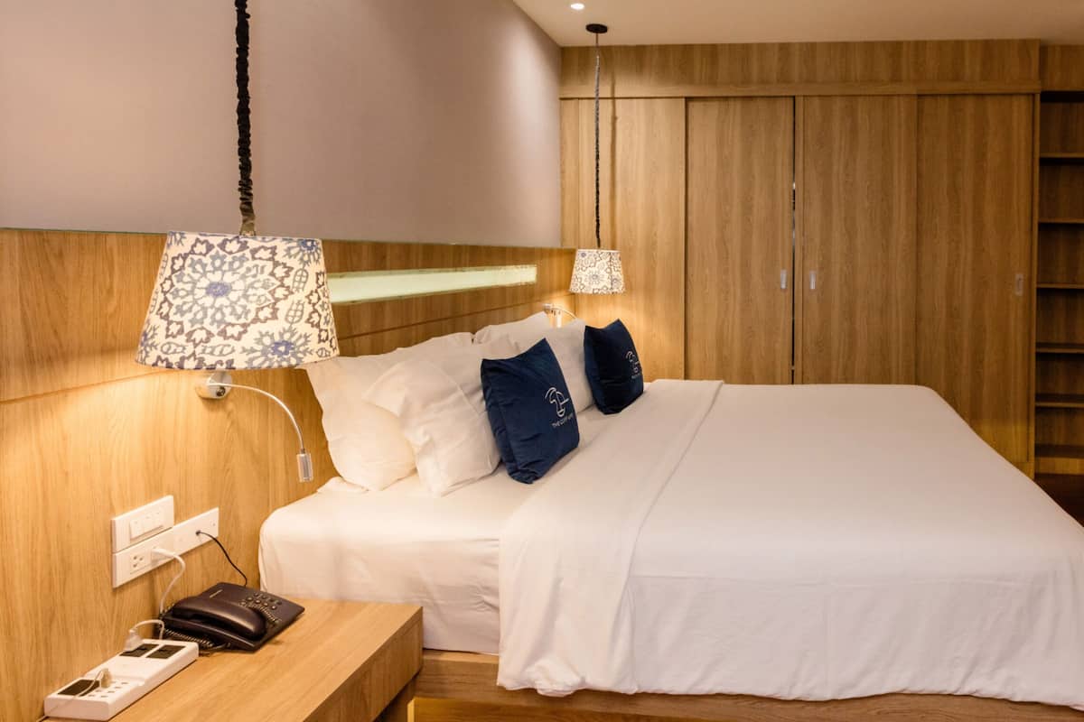 Una cama bien hecha con almohadas blancas y azules, una mesita de noche con lámpara y un armario de madera.