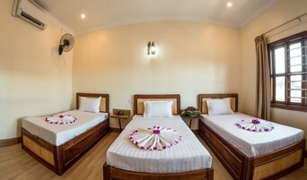 A hotel room with three wooden beds, each with a white bedspread and a floral arrangement. There is a wall-mounted air conditioner and a bedside table with a phone on the left side.