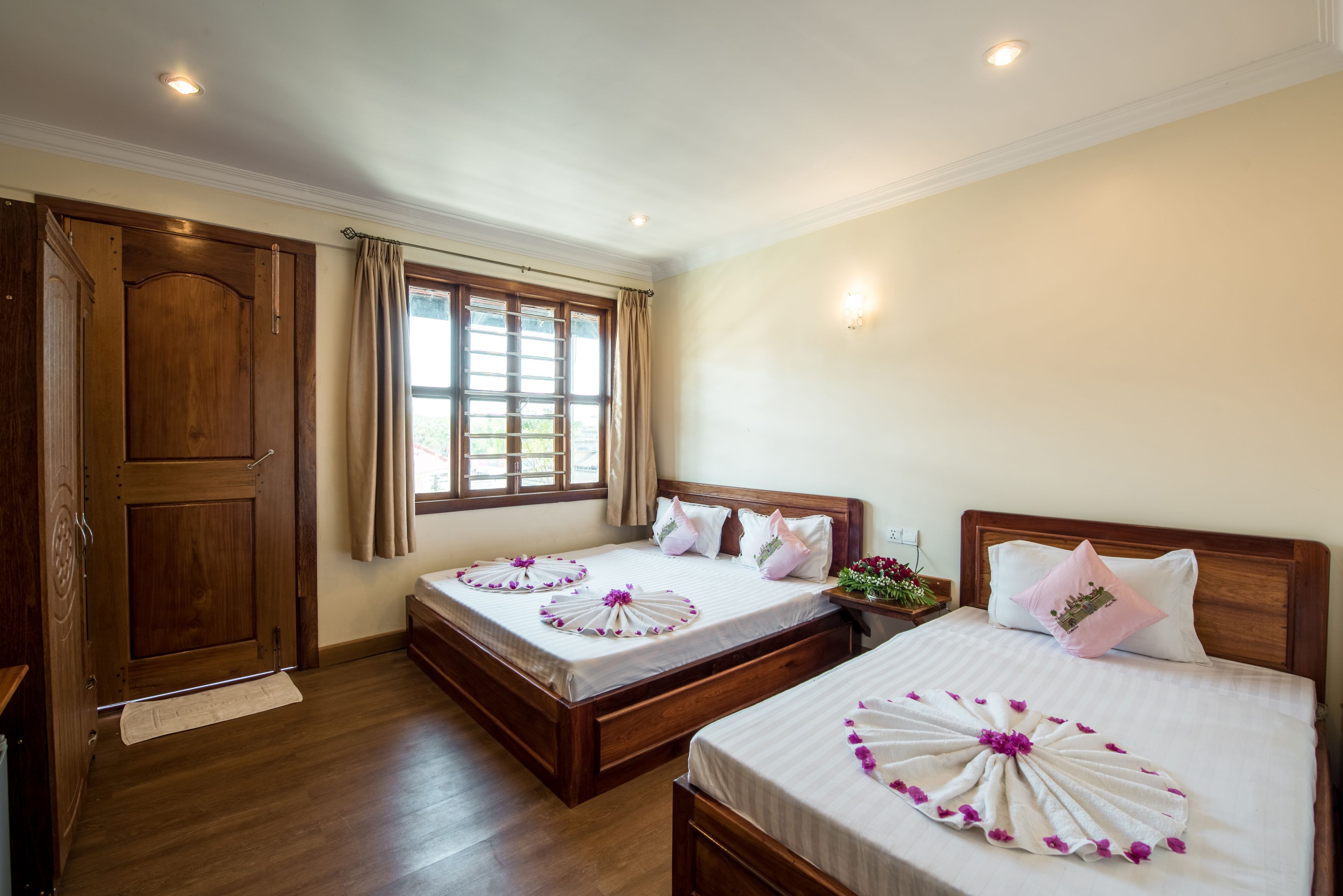 Habitación de hotel con dos camas, piso de madera, un ventanal grande con cortinas y un armario de madera.