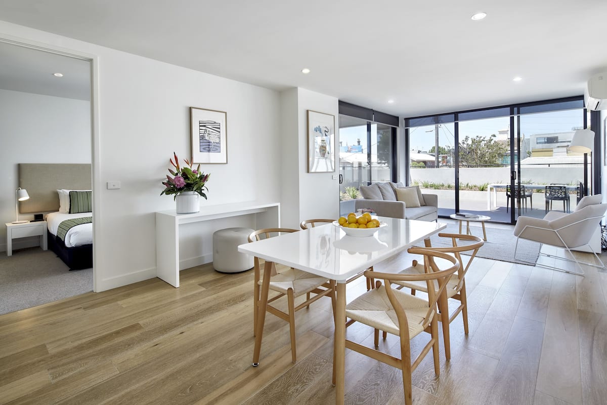 Una sala de estar moderna con una mesa de comedor blanca, sillas de madera y un sofá. Una ventana grande ofrece vistas al exterior.