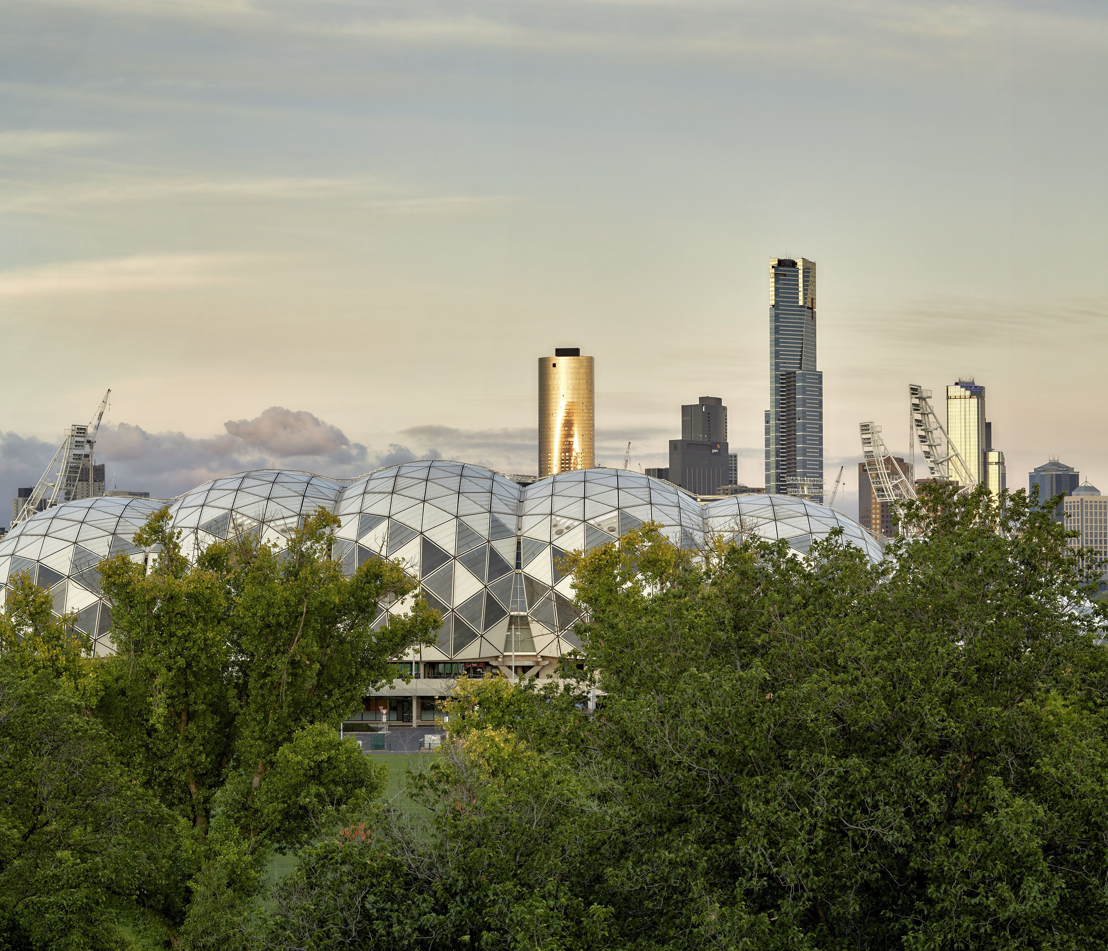 A modern architectural structure with a unique geometric design, surrounded by dense greenery and urban skyscrapers in the background.