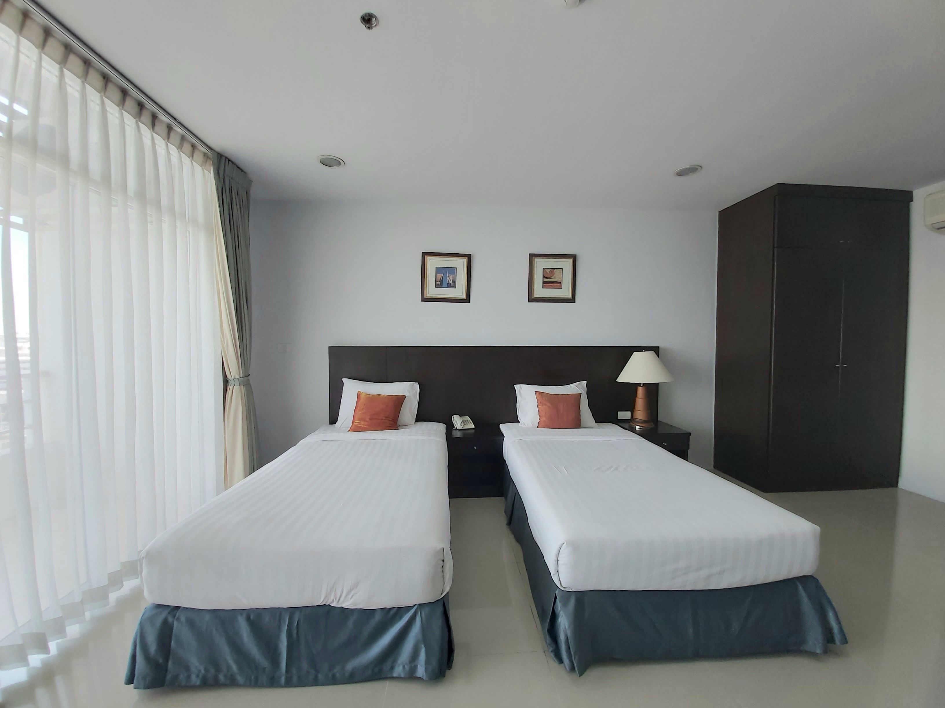 A hotel room with two single beds, a dark wardrobe, a nightstand with a lamp, and two framed pictures on the wall.
