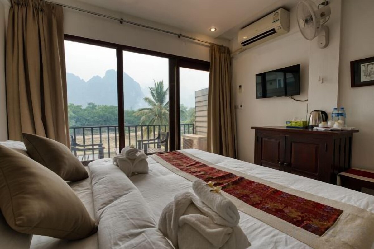 A hotel room with a large bed, a flat-screen TV, a balcony with a view of mountains, and a minibar.