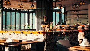A restaurant interior with multiple dining tables, hanging light fixtures, and a large window. - The Elephant Crossing Hotel (Vang Vieng)