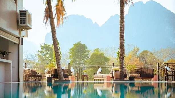 A poolside area with lounge chairs and a view of mountains. - The Elephant Crossing Hotel (Vang Vieng)